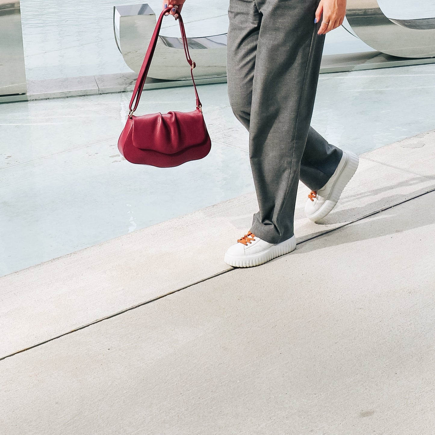 Person holding a red handbag, wearing gray pants and white sneakers, walking on a concrete surface.